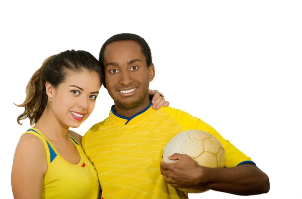 Affascinante coppia interrazziale che indossa camicie da calcio gialle, abbracciando amichevole mentre posa per la palla di tenuta della macchina fotografica, sfondo bianco studio — Foto Stock