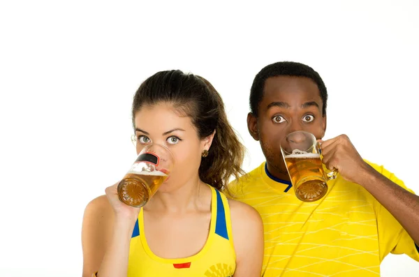 Очаровательная межрасовая пара в жёлтых футбольных футболках, позирует перед камерой, держа пивные очки и улыбаясь, белый студийный фон — стоковое фото