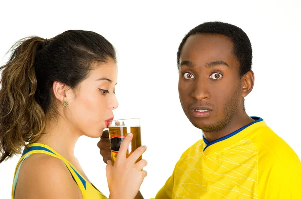 Encantadora pareja interracial con camisas de fútbol amarillo, posando para la cámara mientras la mujer bebe de un vaso de cerveza y el hombre con expresión facial sorprendida, fondo de estudio blanco —  Fotos de Stock