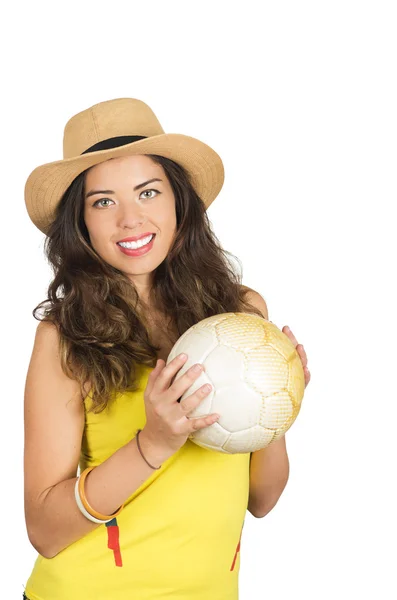 Morena hispana vistiendo camiseta de fútbol amarillo y sombrero, posando para la cámara mientras sostiene la pelota, fondo de estudio blanco — Foto de Stock