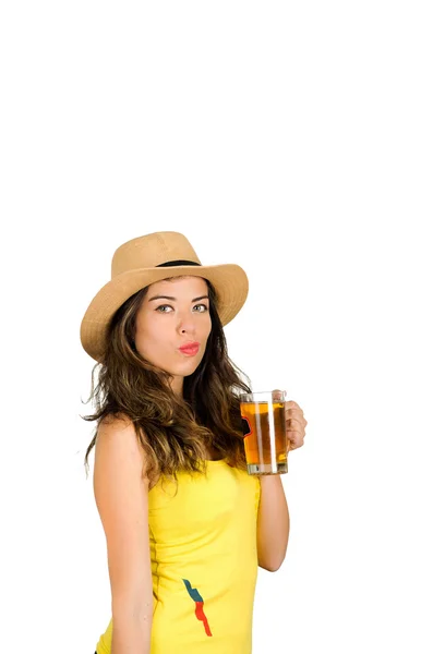 Morena hispana vistiendo camiseta de fútbol amarillo y sombrero, posando para la cámara mientras bebe de un vaso de cerveza, fondo blanco del estudio — Foto de Stock