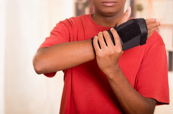 Man in red shirt wearing black wrist brace support on right hand and gripping arm with other — Stock Photo, Image