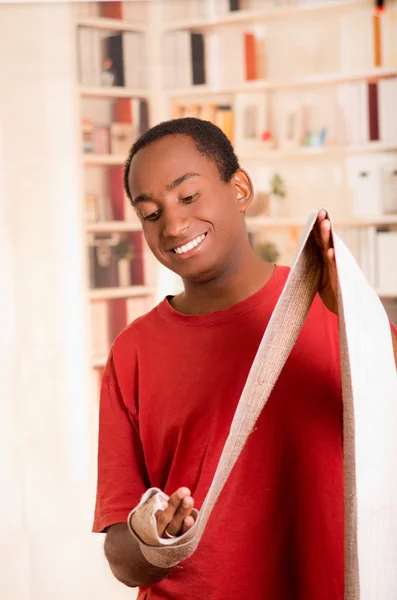 Man in red shirt stretching out bandage preparing wrist support on right hand posing for camera, blurry bookshelves background — Stock Photo, Image