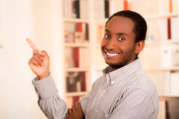 Hombre con ropa casual posando felizmente para la cámara, dedo índice apuntando hacia arriba, fondo de estanterías — Foto de Stock