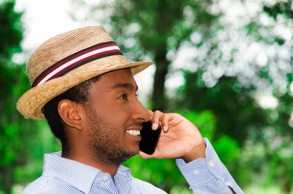 Dobry człowiek patrząc na sobie niebieską koszulę i kapelusz lato rozmowy na telefon komórkowy, podziwiając piękne dni w otoczeniu parku — Zdjęcie stockowe