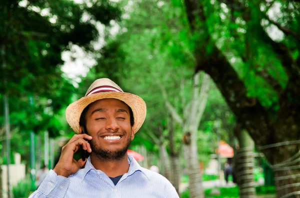 Bell'uomo che indossa camicia blu e cappello estivo parlando sul cellulare mentre si gode una bella giornata nell'ambiente del parco — Foto Stock