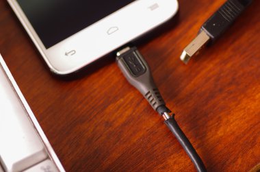 Ahşap yüzey bilgisayar klavye yanında yatan smartphone içine bağlı usb mobil şarj cihazı kablosu kırık, çatlak plastik koruma tel parçası görünen