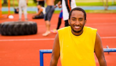 Turuncu atletik yüzeyli açık hava eğitim tesisinde kameraya poz verirken gülümseyen sarı gömlek ve mavi şort giyen adam, bulanık insanlar arka plan