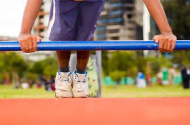 Direğin den sarkan statik mukavemet eksersitler yapan mavi şort giyen adam, turuncu atletik yüzeyli açık hava eğitim tesisi, bulanık insanlar arka plan