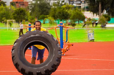 Güç egzersizi sırasında sarı gömlek ve mavi şort giyen adam, turuncu atletik yüzeyli açık hava eğitim tesisi, yeşil ağaçlar arka plan