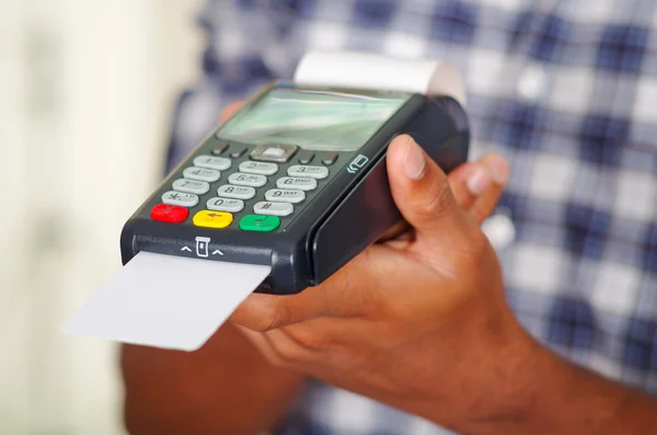Mann in blau-weißem Hemd mit quadratischem Muster hält Kreditkartenzahlungsterminal vor Kamera hoch, Nahaufnahme — Stockfoto