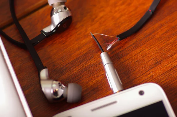 Broken audio headphones cable connected into smartphone lying on wooden surface, part of wire exposed through crack in plastic protection — Stock Photo, Image