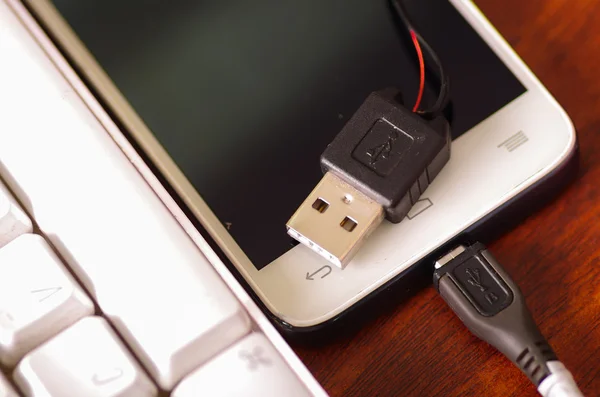 Gebrochenes USB-Ladekabel für Mobiltelefone, das mit dem Smartphone verbunden ist, liegt auf einer Holzoberfläche neben der Computertastatur, ein Teil des Drahtes ist durch Risse im Kunststoffschutz freigelegt — Stockfoto