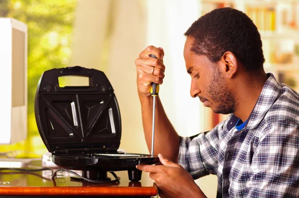 Hombre sentado junto al escritorio reparando sandwichera usando destornillador, expresiones faciales serias mientras trabaja — Foto de Stock