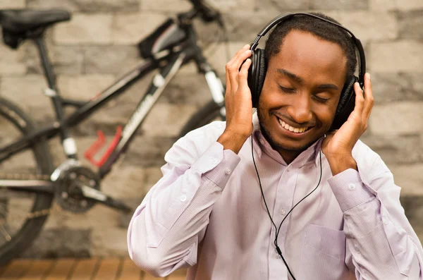 Man wearing white red business shirt sitting down with headphones on, enjoying music while smiling happily, bicycle standing behind leaning against grey brick wall — Stock Photo, Image
