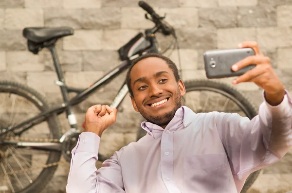 Hombre con camisa blanca roja de negocios sentado, sosteniendo el teléfono móvil tomando foto selfie, sonriendo y posando, bicicleta de pie detrás apoyado contra la pared de ladrillo gris — Foto de Stock