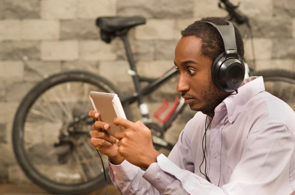 Man wearing white red business shirt sitting down, headphones on, looking at tablet screen with intense stare, bicycle standing behind against grey brick wall — Stock Photo, Image