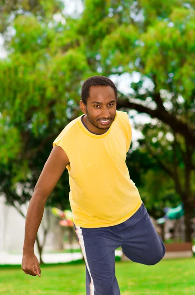 Uomo che indossa camicia gialla e pantaloncini blu stretching gambe utilizzando le mani nel parco circondato da alberi di erba verde, concetto di allenamento — Foto Stock