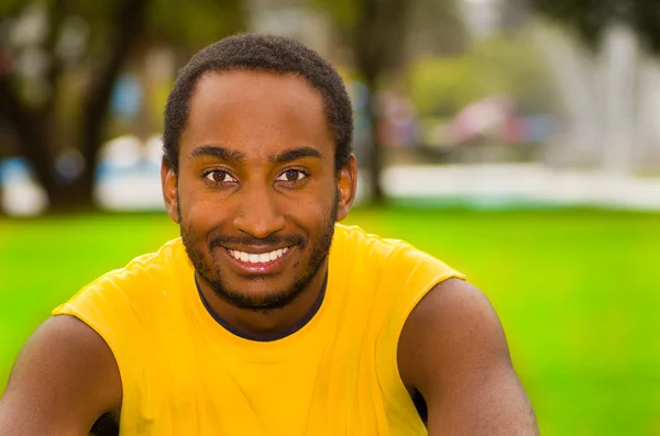 Uomo con camicia gialla seduto nel parco circondato da erba verde e alberi, espressione del viso felice, concetto di allenamento — Foto Stock