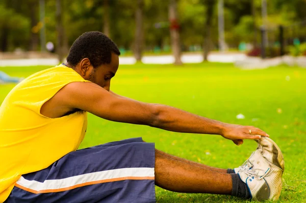 Hombre con camisa amarilla y pantalones cortos azules sentado en la hierba verde estiramiento tocando zapatos con las manos, concepto de entrenamiento — Foto de Stock