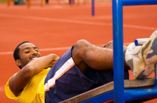 Hombre con camisa amarilla y pantalones cortos azules acostado en tablón de entrenamiento de madera haciendo situps sonriente, fondo de superficie atlética naranja — Foto de Stock