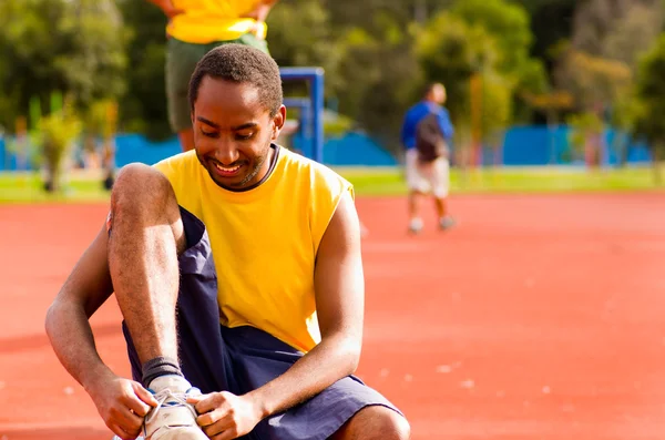 Man wearing yellow shirt and blue shorts sitting down tying shoe laces at outdoors training facility with orange athletic surface, blurry people background