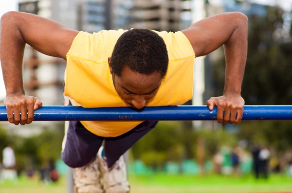 Hombre con camisa amarilla y pantalones cortos azules haciendo ejercicios de fuerza estática colgando del poste, instalación de entrenamiento al aire libre con superficie atlética naranja, fondo borroso de la gente — Foto de Stock