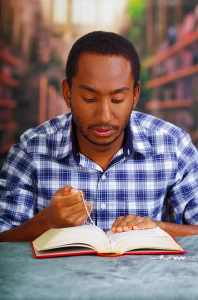 Uomo religioso seduto tenendo il rosario, pregando e leggendo dal libro aperto sulla scrivania di fronte, concetto di religione — Foto Stock