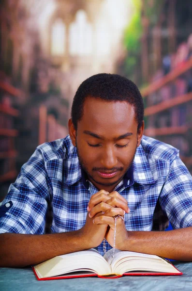 Uomo religioso seduto tenendo il rosario, pregando e leggendo dal libro aperto sulla scrivania di fronte, concetto di religione — Foto Stock