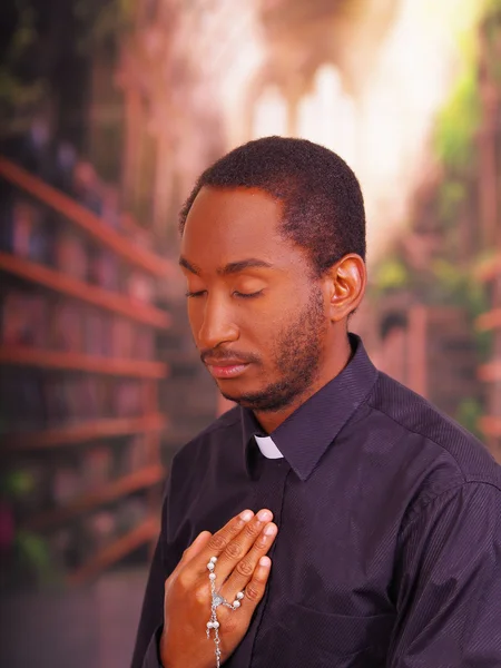 Sacerdote cattolico che indossa la tradizionale camicia del colletto clericale in piedi eseguendo segno della croce e tenendo in mano il rosario, concetto di religione — Foto Stock