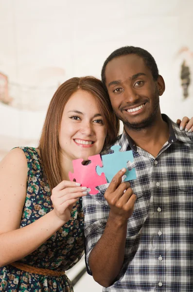 Interracial charming couple embracing friendly, holding up large puzzle pieces and happily interacting having fun, blurry studio background — Stock Photo, Image