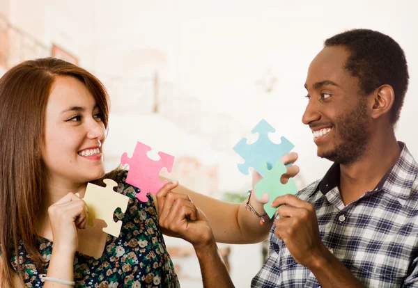 Interracial casal encantador segurando grandes peças de quebra-cabeça e felizmente interagindo se divertindo, embaçado estúdio fundo — Fotografia de Stock