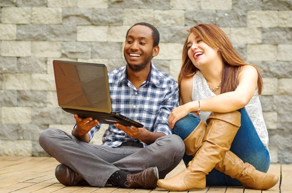 Ein charmantes Paar in lässiger Kleidung sitzt auf einer Holzfläche und schaut gemeinsam auf den Laptop vor einer grauen Backsteinwand. — Stockfoto