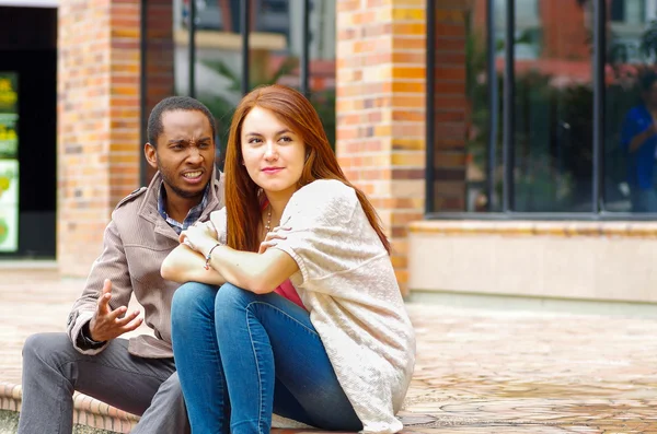 Interracial happy charming couple sitting on steps in front of building interacting and smiling for camera — Stock Photo, Image