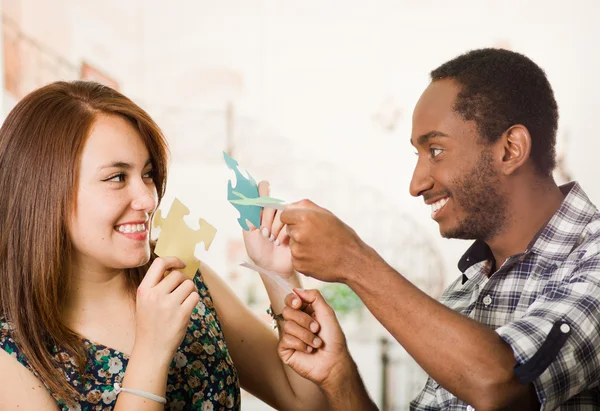 Interracial casal encantador segurando grandes peças de quebra-cabeça e felizmente interagindo se divertindo, embaçado estúdio fundo — Fotografia de Stock