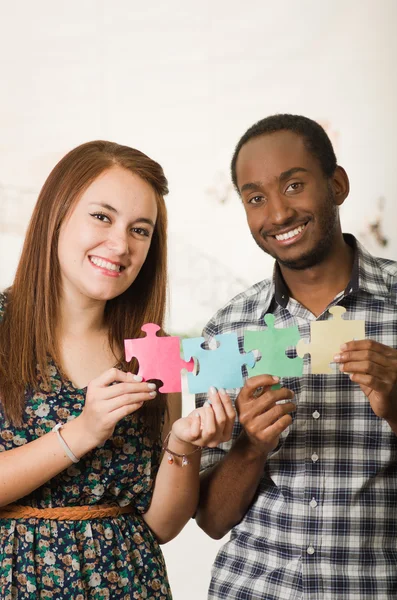 Interracial charmantes Paar in lässiger Kleidung hält große Puzzleteile hoch und interagiert fröhlich, weißer Studiohintergrund — Stockfoto