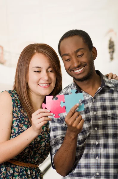 Interracial charming couple embracing friendly, holding up large puzzle pieces and happily interacting having fun, blurry studio background — Stock Photo, Image