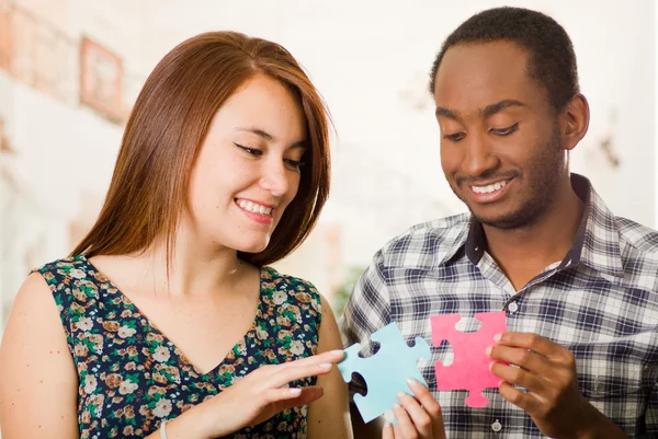 Interracial charming couple holding up large puzzle pieces and happily interacting having fun, blurry studio background — Stock Photo, Image