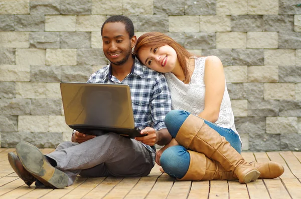 Ein charmantes Paar in lässiger Kleidung sitzt auf einer Holzfläche und schaut gemeinsam auf den Laptop vor einer grauen Backsteinwand. — Stockfoto