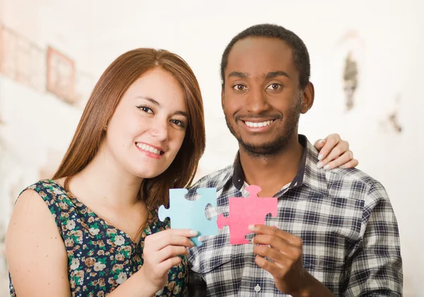 Interracial charming couple embracing friendly, holding up large puzzle pieces and happily interacting having fun, blurry studio background — Stock Photo, Image