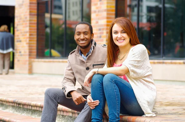 Межрасовая счастливая очаровательная пара, сидящая на ступеньках перед зданием, взаимодействующая и улыбающаяся на камеру — стоковое фото