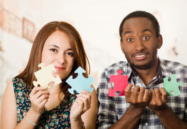 Interracial charming couple holding up large puzzle pieces and happily interacting having fun, blurry studio background — Stock Photo, Image