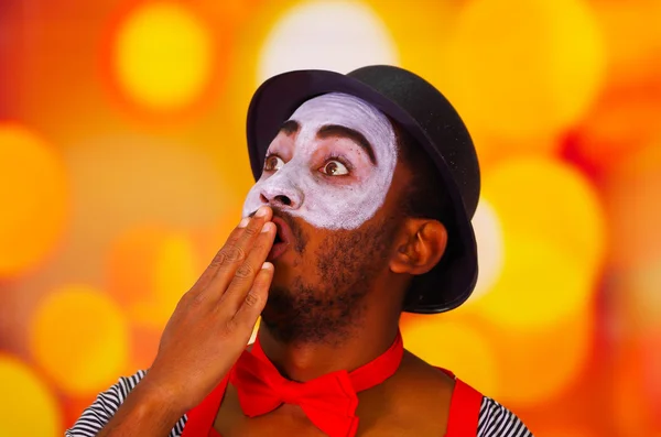 Headshot pantomima uomo con vernice facciale in posa per la fotocamera utilizzando le mani per coprire la bocca, luci sfocate sfondo — Foto Stock