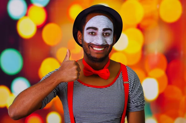Homem pantomima com pintura facial posando para câmera interagindo dando polegares para cima sorrindo, luzes embaçadas fundo — Fotografia de Stock
