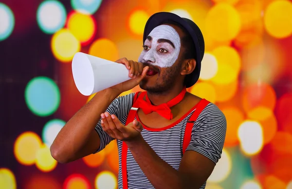 Homem pantomima com pintura facial posando para câmera segurando chifre de sopro, luzes embaçadas fundo — Fotografia de Stock