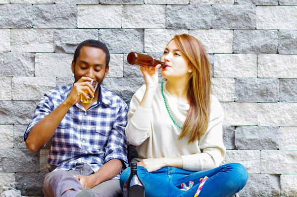 Casal inter-racial vestindo roupas casuais sentado na parede de tijolo cinza desfrutando de algumas bebidas e companhia uns dos outros — Fotografia de Stock