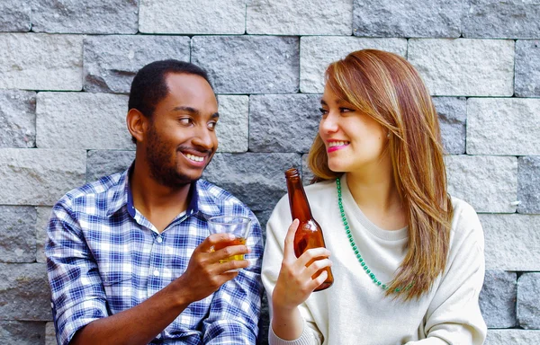 Casal inter-racial vestindo roupas casuais sentado na parede de tijolo cinza desfrutando de algumas bebidas e companhia uns dos outros — Fotografia de Stock