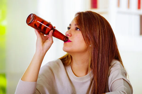 Mujer atractiva vistiendo suéter blanco sentado junto al mostrador de bar bebiendo de la botella de cerveza, borracho expresión facial deprimida, concepto alcohólico — Foto de Stock