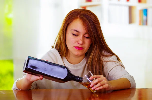 Aantrekkelijke vrouw dragen witte trui zit door bar teller gieten drankje uit fles in een shot glas, dronken depressief gelaatsuitdrukking, alcoholische concept — Stockfoto