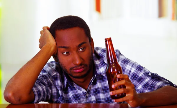 Beau homme portant une chemise bleue blanche assise à côté d'un comptoir de bar couché sur un bureau tenant une bouteille de bière brune, expression faciale déprimée ivre, concept alcoolique — Photo
